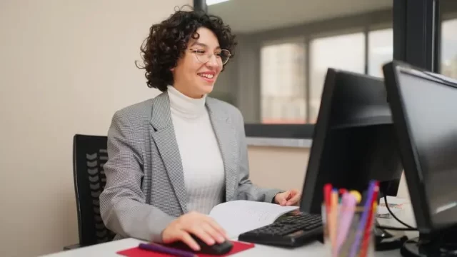 A person with curly hair and glasses is sitting at a desk in an office, smiling while using a computer mouse. An open laptop and colorful pens underscore their dedication to personal development. They're wearing a gray blazer over a white turtleneck, embodying the importance of investing in yourself.