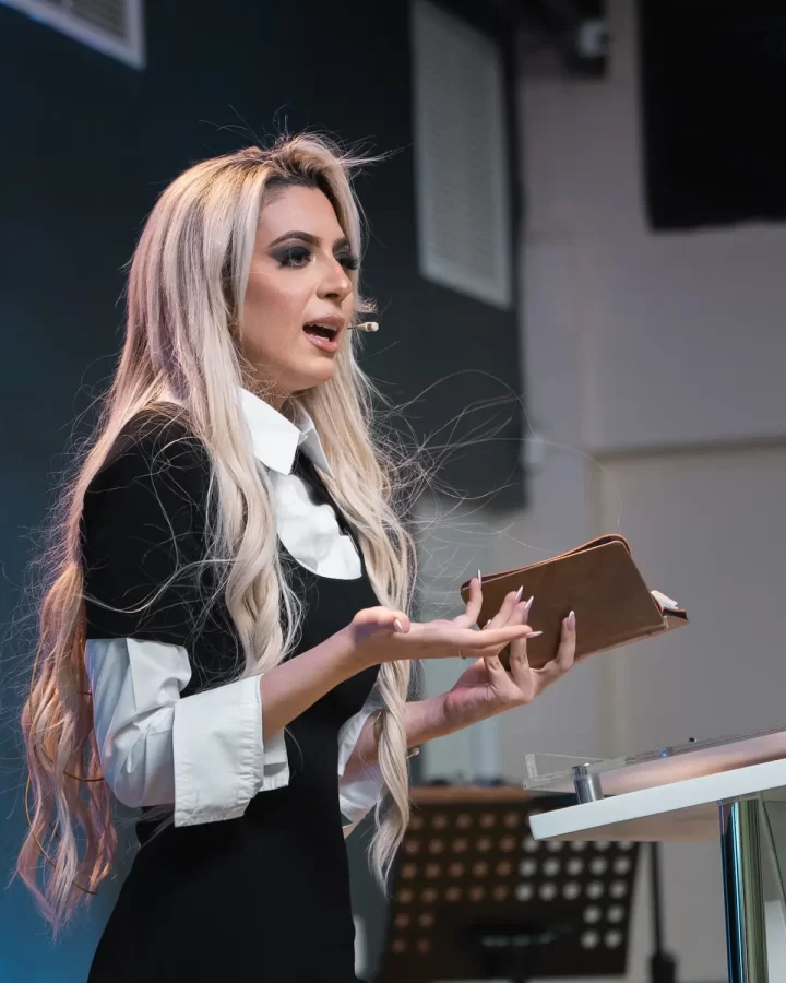 A woman with long blonde hair, wearing a black and white outfit, speaks passionately about mental patterns while holding an open book at a podium. She is in a room with a microphone headset.