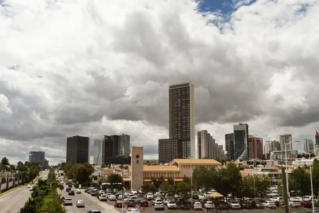 City skyline with modern high-rise buildings under a cloudy sky. A busy road with cars runs through the foreground, flanked by trees. A historic clock tower and a traditional building are visible among the contemporary structures.