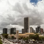 City skyline with modern high-rise buildings under a cloudy sky. A busy road with cars runs through the foreground, flanked by trees. A historic clock tower and a traditional building are visible among the contemporary structures.