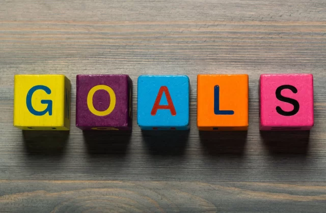 Colorful wooden blocks spelling "GOALS" are arranged in a row on a wooden surface. Each block has a different color: yellow, purple, blue, orange, and pink, with black and red letters.