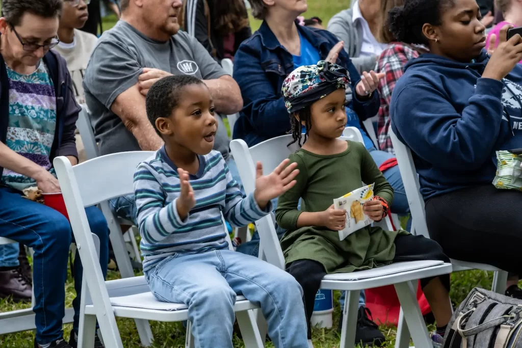 Two young children sit on white chairs in an outdoor setting. The boy on the left is enthusiastically clapping, looking ahead with joy, while the girl beside him wears a colorful helmet and holds a book. Several adults are seated in the background.