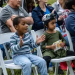 Dos niños pequeños están sentados en sillas blancas al aire libre entre un grupo de personas. El niño de la izquierda, que lleva una camiseta a rayas, aplaude. La niña de la derecha, que lleva un vestido verde y un casco, sostiene una bolsa con merienda. Hay otros sentados detrás de ellos.