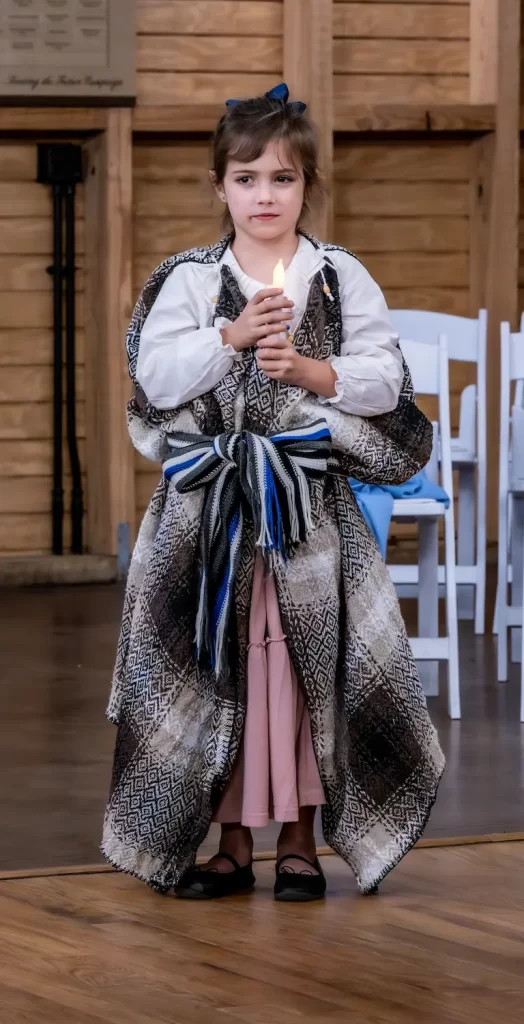 A young girl stands indoors, looking ahead while holding a lit candle. She wears a patterned robe over a white shirt and pink skirt, with a blue ribbon in her hair. White chairs and a wooden wall are in the background, creating an ambiance of quiet reflection.