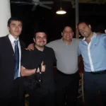 Four men are standing together, smiling, and gesturing thumbs up. They are in a casual setting, possibly at a party or gathering. The man on the left is wearing a suit, while the others are dressed in more casual attire.