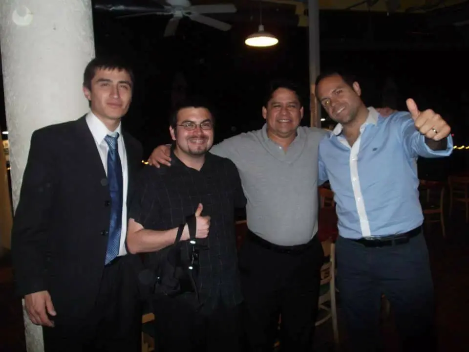 Four men are standing together, smiling, and gesturing thumbs up. They are in a casual setting, possibly at a party or gathering. The man on the left is wearing a suit, while the others are dressed in more casual attire.