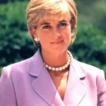 Una mujer de pelo corto y rubio, con una chaqueta color lavanda, un collar de perlas y pendientes de perlas, está parada al aire libre frente a un jardín. Tiene una expresión amable en su rostro.