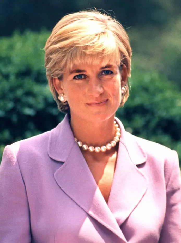 Woman with short blonde hair wears a pastel purple suit and pearl necklace, standing outdoors with greenery in the background. She gazes at the camera with a slight smile, evoking timeless elegance reminiscent of Princess Diana's grace within the Royal Family.