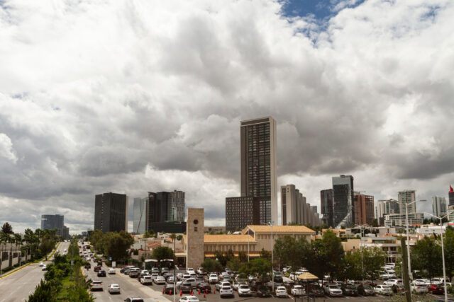 Paisaje urbano con modernos rascacielos bajo un cielo nublado. En primer plano se puede ver un aparcamiento lleno de vehículos y un edificio histórico. A la izquierda, una carretera ancha con poco tráfico discurre junto a la vegetación.