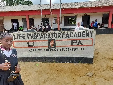 A young girl stands in front of a sign for Life Preparatory Academy, which reads "Motto: We Prepare Students for Life." A school building with red accents and people sitting nearby is visible in the background.