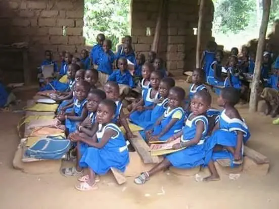 Children in blue uniforms sit closely together on wooden benches in a rustic classroom. The room is made of unfinished bricks, and several children hold books. Natural light enters through large openings.