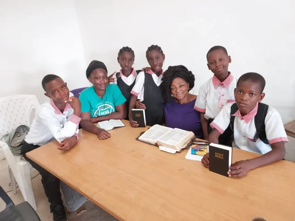 A group of five students in school uniforms and three adults gather around a table with open books, including a Bible, smiling at the camera. The background is a plain white wall.