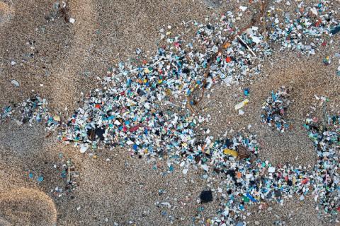 Vista aérea de una playa de arena con coloridos trozos de desechos plásticos dispersos en la superficie, creando un contraste ambiental contra el fondo de arena natural.