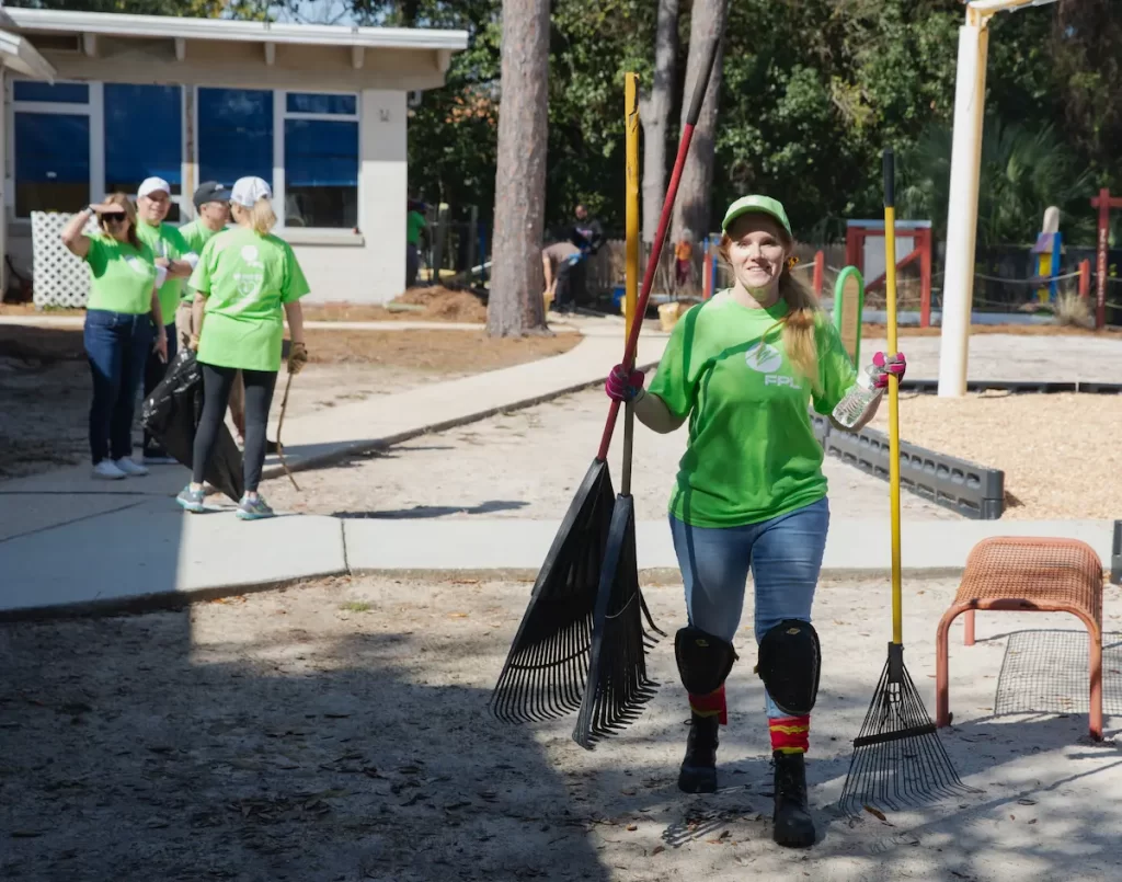 A woman in a green t-shirt and hat holds two rakes and a plastic bottle, actively contributing to the Beautify initiative. In the background, FPL Volunteers in similar green attire help clean up litter near a building and playground area in the park.