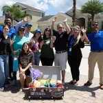 A group of people smiling and raising their hands poses around a basket filled with goodies. They're standing on a paved driveway in front of suburban homes. A partly cloudy sky and palm trees are in the background.