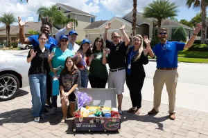 A group of people smiling and raising their hands poses around a basket filled with goodies. They're standing on a paved driveway in front of suburban homes. A partly cloudy sky and palm trees are in the background.