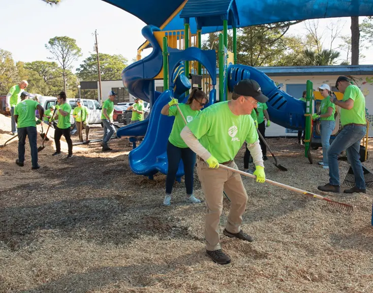 A group of FPL Volunteers wearing green shirts and gloves works together to beautify the playground area at The Arc Gateway. Some use rakes and shovels to spread mulch, while a large blue slide stands in the background alongside trees and a building.