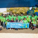 Un grupo de personas con camisetas verdes iguales y gafas de sol posa con una pancarta que dice "Poder para Cuidar: Apoyando a las comunidades donde vivimos y trabajamos". Se encuentran frente a un colorido parque infantil bajo una gran estructura de sombra.