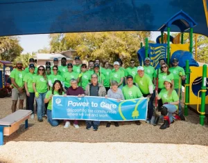 Un grupo de personas con camisetas verdes iguales y gafas de sol posa con una pancarta que dice "Poder para Cuidar: Apoyando a las comunidades donde vivimos y trabajamos". Se encuentran frente a un colorido parque infantil bajo una gran estructura de sombra.