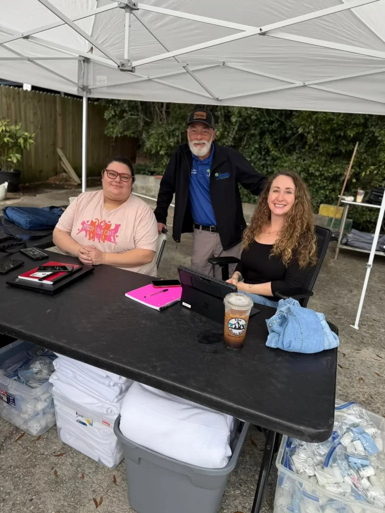 Three people enjoy an outdoor event under a white canopy in Okaloosa. Two women, one in a pink shirt and the other in a black top, sit at a table with devices and drinks. A man stands behind them wearing a cap and jacket, while supplies related to homelessness are visible under the table.