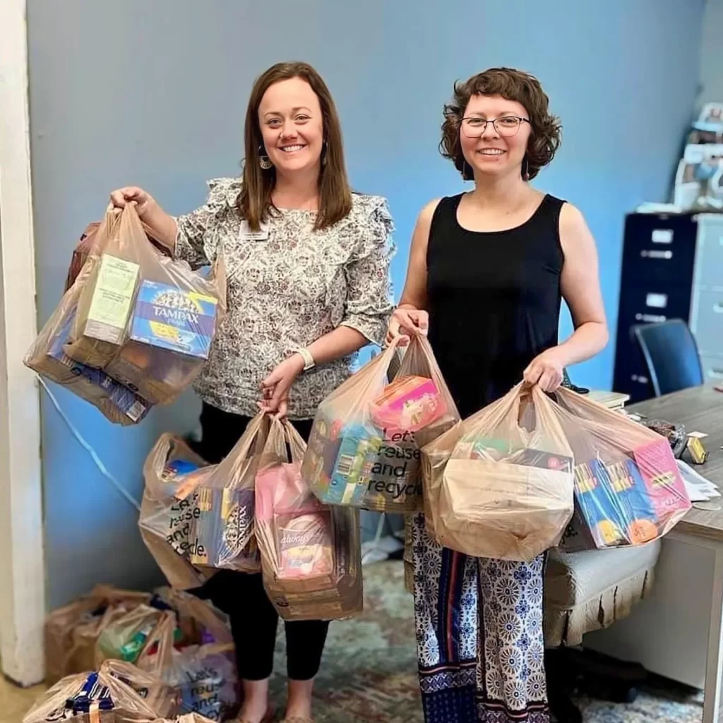 Two women, affiliated with the Housing Alliance, smile as they hold several plastic bags filled with assorted groceries and household items in an office setting. A desk and filing cabinet are visible in the background, highlighting their efforts to combat homelessness in Okaloosa Counties.