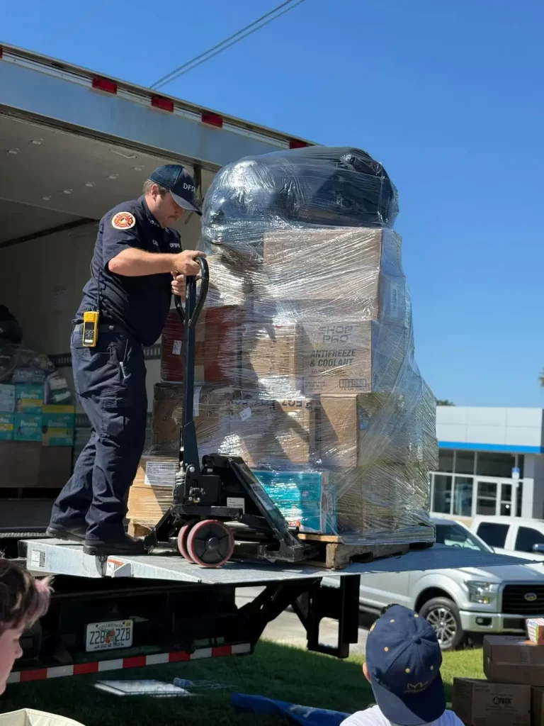 A worker in uniform and cap expertly maneuvers a pallet jack to unload shrink-wrapped boxes from a truck, while the Housing Alliance team observes. The scene unfolds outdoors on a sunny day in Okaloosa Walton Counties.