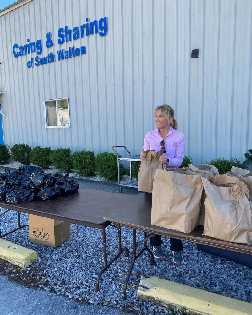 A person in a pink shirt stands behind a table with brown and black bags outside a building labeled "Caring & Sharing of South Walton." They are smiling and holding a bag, embodying the support provided to tackle homelessness in Okaloosa and Walton Counties.