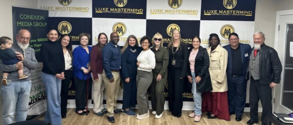 A diverse group of people, standing in a row, smile for a photo against a backdrop with logos reading "Luxe Mastermind" and "Conexion Media Group." A man holding a child stands proudly on the left, embodying the spirit of Luxe collaboration.