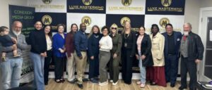 A diverse group of people, standing in a row, smile for a photo against a backdrop with logos reading "Luxe Mastermind" and "Conexion Media Group." A man holding a child stands proudly on the left, embodying the spirit of Luxe collaboration.