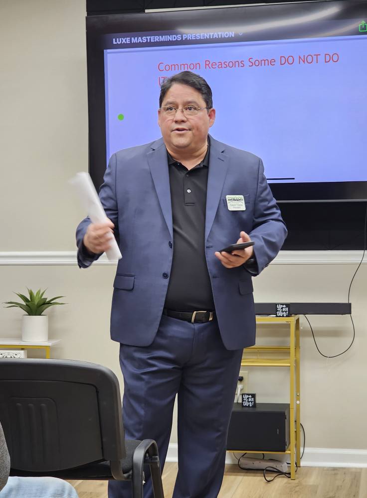 A person in a suit stands in front of a presentation screen, exuding a luxe aura while holding a piece of paper and a phone. The screen displays the words "Common Reasons Some DO NOT DO." Nearby, an indoor plant decorates the shelf, adding to the mastermind's polished setting.