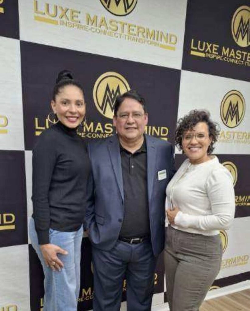 Three people stand smiling in front of a "Luxe Mastermind" backdrop. The two women flank a man in a suit, embodying the luxury and collaboration that define this professional gathering. Their close stance suggests a welcoming and insightful environment.