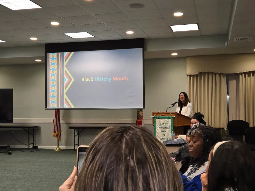 A woman stands at a podium in a conference room, addressing an audience. A screen displays "Black History Month" with a colorful pattern on the left. Attendees are seated, and an American flag is visible in the background.