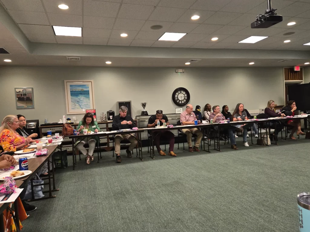 A group of people sitting around three sides of a rectangular table in a conference room, engaged in writing and looking at documents. The room has a painting, a clock on the wall, and a projector on the ceiling.