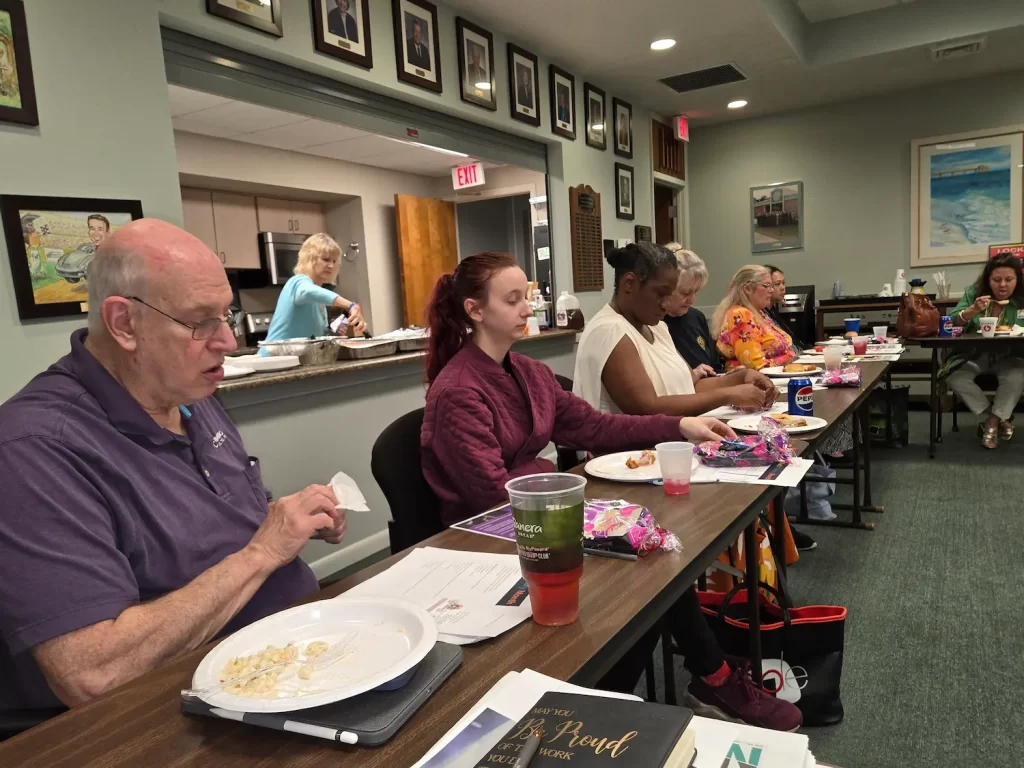 A group of people sits at a table in a meeting room with food and drinks. They are engaged in eating and conversation. The room has photos on the wall and a serving area in the background.