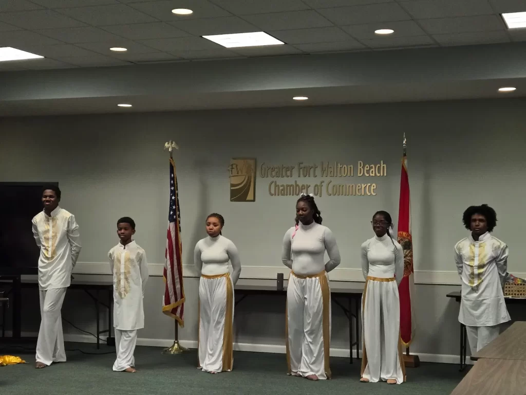 A group of seven people stand in a room, wearing white outfits with gold accents. They are in front of a wall with a "Greater Fort Walton Beach Chamber of Commerce" sign and two flags. The room has a conference setting.