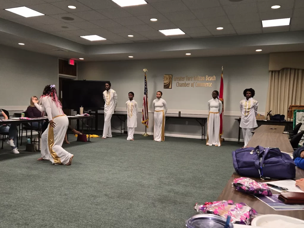 A group of five dancers in white outfits with gold accents perform in a meeting room. An audience sits around tables. An American flag and a chamber of commerce sign are displayed in the background.