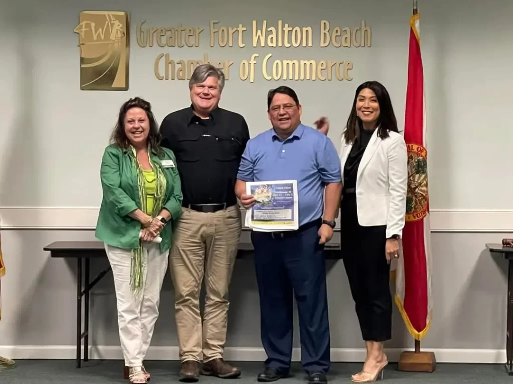 Cuatro personas están de pie sonriendo frente a una pared con el logotipo de la Cámara de Comercio de Greater Fort Walton Beach. Una persona sostiene un certificado. El grupo incluye dos mujeres y dos hombres, con banderas a ambos lados.