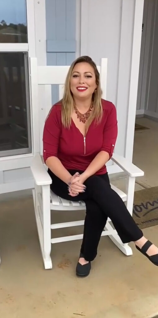 A person with long blonde hair is sitting on a white rocking chair on a porch, embodying the essence of "My Journey." They wear a red blouse, black pants, and shoes, complemented by a red necklace. Their smile glows as their hands are clasped in calm reflection.