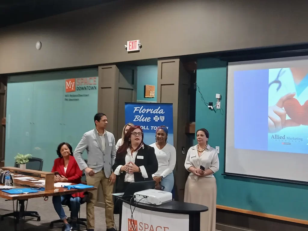 Five people are standing at the front of a room next to a presentation screen showing a health-related slide. A banner reads "Florida Blue." A woman is sitting at a table with papers. The room has a teal accent wall and wood decor.