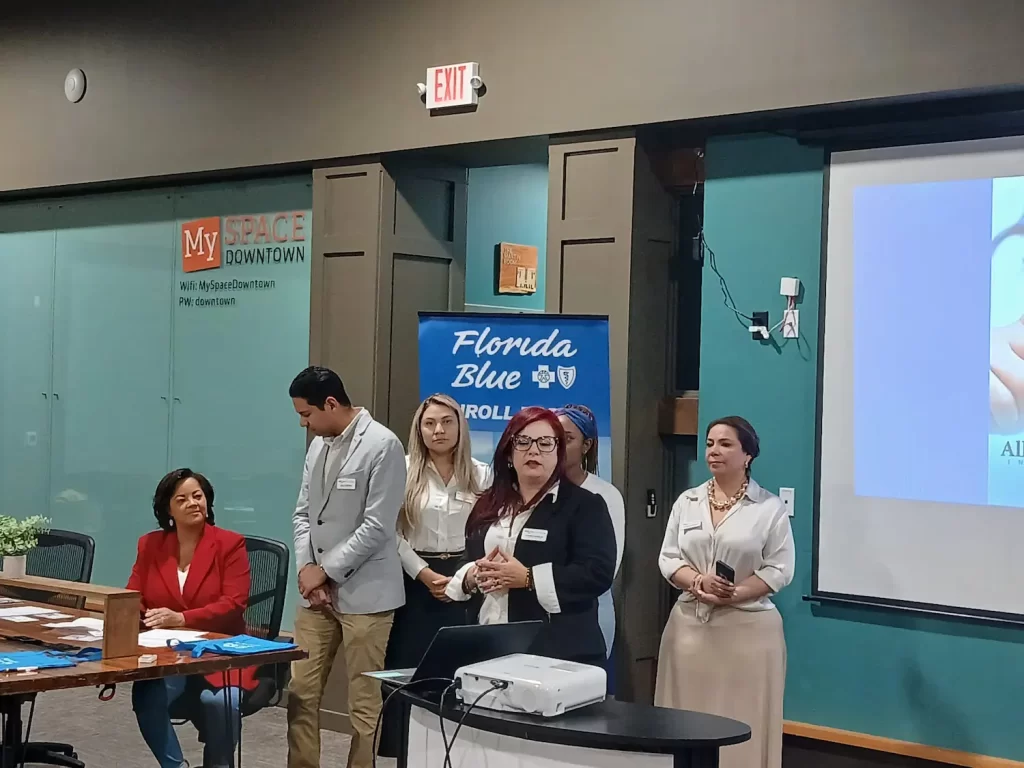 A group of people stands at the front of a room near a projector, participating in a presentation. A blue banner in the background reads "Florida Blue." A woman in red sits at a table with blue items spread on it.