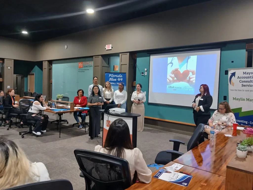 Un grupo de personas está haciendo una presentación en una sala de conferencias. Una persona habla en un podio y otras permanecen de pie cerca. En la pantalla se proyecta una diapositiva que muestra un corazón. Los asistentes están sentados en una mesa larga y se muestran varias pancartas.