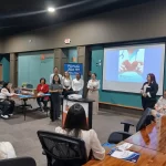 Un grupo de personas está haciendo una presentación en una sala de conferencias. Una persona habla en un podio y otras permanecen de pie cerca. En la pantalla se proyecta una diapositiva que muestra un corazón. Los asistentes están sentados en una mesa larga y se muestran varias pancartas.