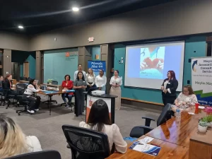 A group of women stands at the front of a conference room, next to a screen displaying a presentation titled "Allied Access Care." Attendees sit around a large table, and banners with business logos are visible in the room.