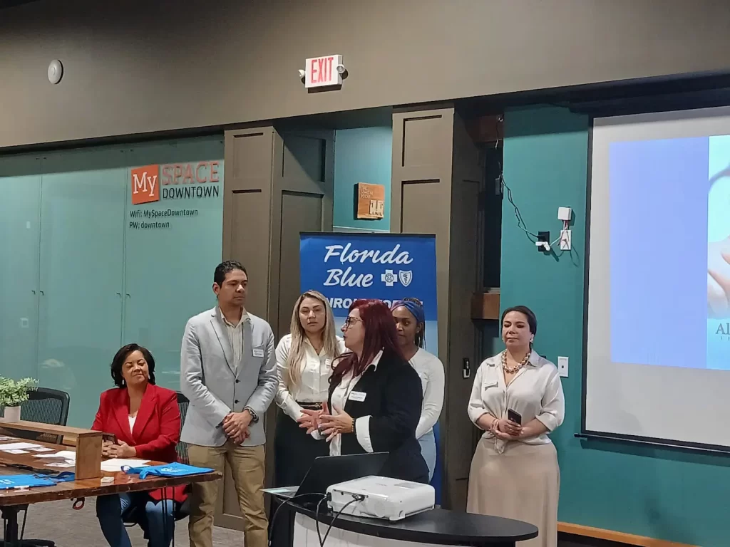 A group of people stand in front of a presentation screen and a banner that reads "Florida Blue." One person is speaking, while others listen beside them. A woman sits at a table with papers, and a laptop is open. They are in a room with a glass wall.