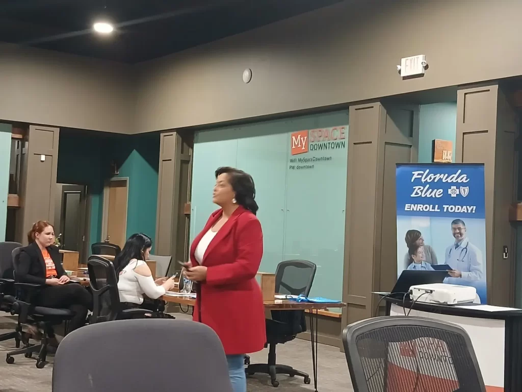 A person in a red coat stands speaking in a room with desks and seated people. A banner reads "Florida Blue. Enroll Today!" with images of two smiling people. The setting appears to be a conference or meeting space.