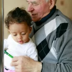 Un hombre mayor con un suéter gris y negro sostiene a un niño pequeño en su regazo. El niño lleva una camisa blanca y pantalones rosas con personajes de dibujos animados y mira un bolígrafo verde que el hombre sostiene. Están en un interior con un fondo neutro.