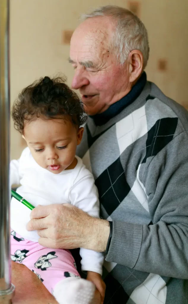 An elderly man with white hair tenderly holds a small child on his lap. The child, with curly hair, curiously examines a green pen in the older man's hand. Clad in a gray argyle sweater, the man complements the child's white shirt and pink pants.