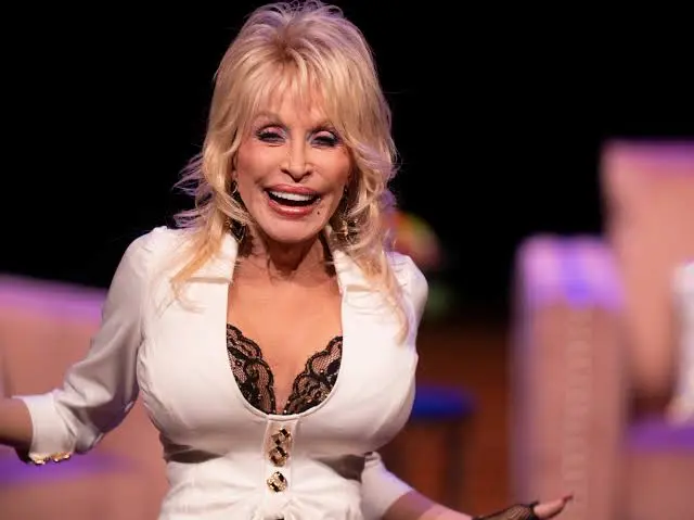 A person with long blonde hair, reminiscent of Dolly Parton, and a big smile is wearing a white top with a black lace trim. They are standing in front of a softly blurred background that features pink and purple hues.