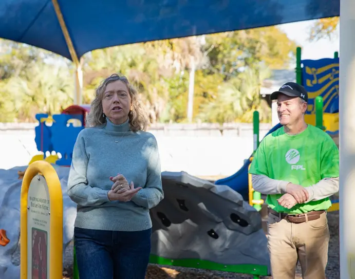 Two people stand in a playground under a blue sunshade, part of an FPL Volunteers' effort to beautify the space. The person on the left is speaking, wearing a gray sweater and jeans. The person on the right wears a green shirt and cap, smiling with hands clasped. Playground equipment is visible in the background.