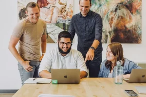 Four people gather around a table with laptops, notebooks, and a water bottle. They are smiling and engaged in discussion, confidently showcasing their work. A colorful painting hangs on the wall behind them, highlighting their accomplishments.